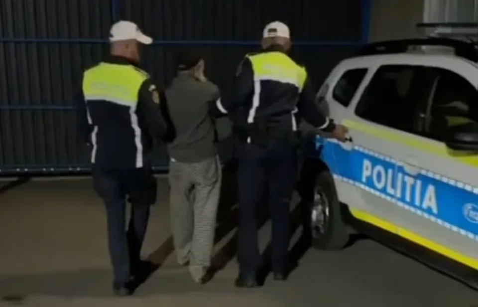 Doi polițiști în uniformă cu benzi reflectorizante conduc un bărbat într-o direcție lângă un autoturism de poliție albastru și alb.
