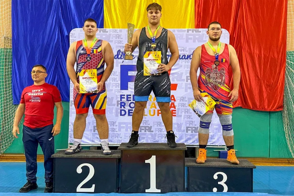 Trei bărbați în costume de wrestling stau pe un podium de premiere în fața unui fundal colorat.