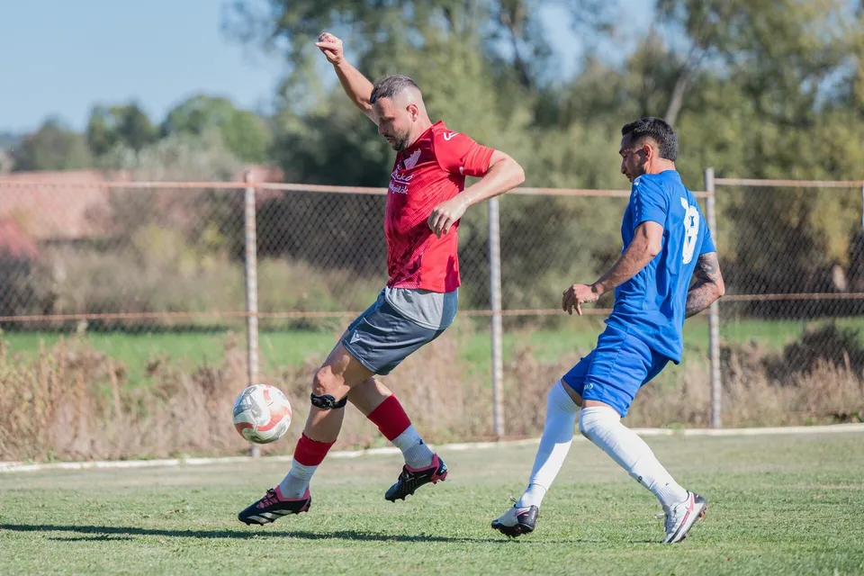 Un jucător în echipament roșu și gri lovește un fotbal pe un teren de fotbal, în timp ce un alt jucător în albastru îl urmărește.
