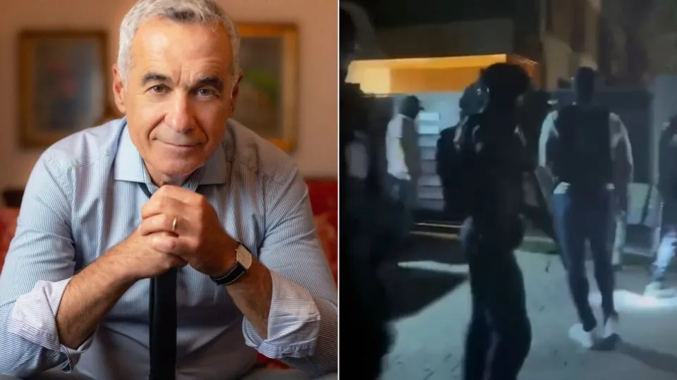 Un bărbat cu părul cărunt stă la o masă, iar în partea dreaptă se observă siluete de persoane în întuneric pe un drum.