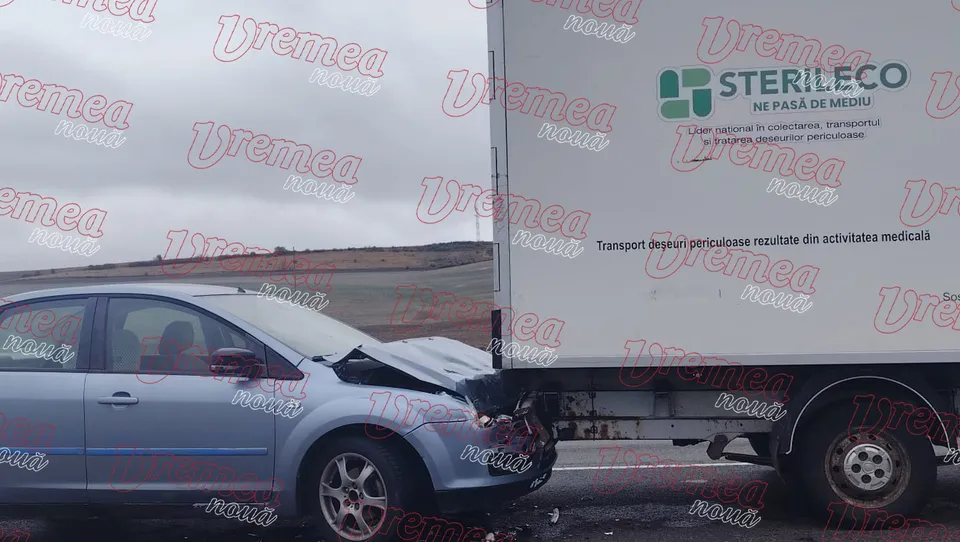 Un autoturism de culoare albastră este colțuit de partea din spate de un camion alb cu inscripții pe parte.
