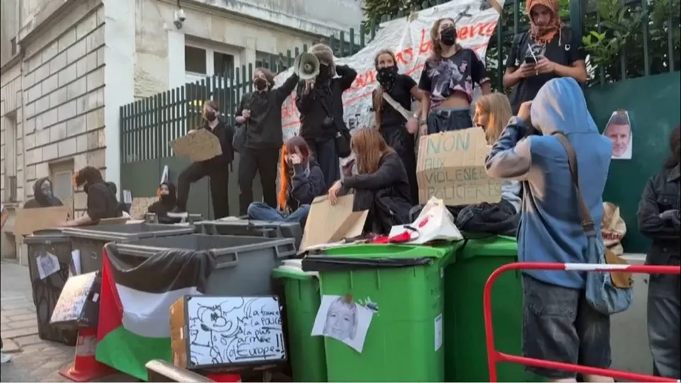 Un grup de persoane stă pe un gard și lângă containere de gunoi, purtând haine în culori închise și mască, într-un spațiu exterior.