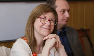O femeie cu ochelari roșii și părul castaniu stă cu mâinile subțacând chinul, zâmbind ușor, în timp ce un bărbat stă în fundal.