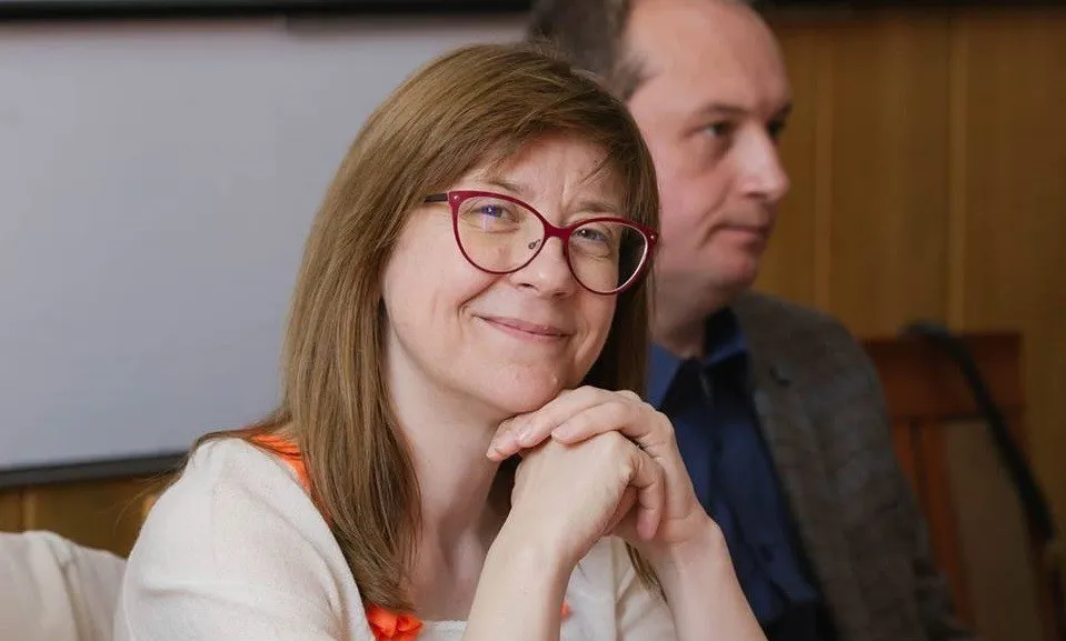 O femeie cu ochelari roșii și părul castaniu stă cu mâinile subțacând chinul, zâmbind ușor, în timp ce un bărbat stă în fundal.