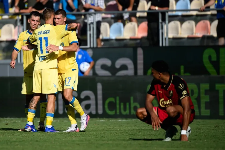 Trei jucători de fotbal în echipament galben se îmbrățișează pe teren, în timp ce un jucător în echipament roșu și negru stă ghemuit pe iarbă.