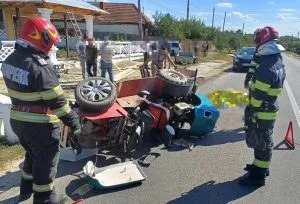 Două persoane în uniforme de pompier stau lângă un autoturism răsturnat pe o stradă asfaltată lângă un motocultor.
