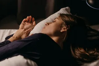 O femeie cu părul lung doarme pe o pernă albă, având mâinile ridicate lângă față într-un mediu întunecat.