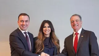 Trei persoane zâmbesc, un bărbat în costum în stânga strâng mâna unei femei cu părul lung, aflată între un alt bărbat în costum.