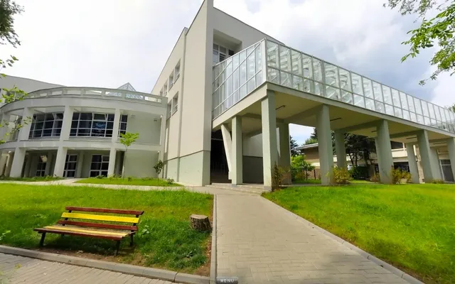 O clădire modernă cu o pasarelă acoperită de sticlă este situată lângă un teren de gazon verde cu o bancă de lemn colorată.