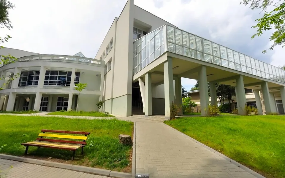 O clădire modernă cu o pasarelă acoperită de sticlă este situată lângă un teren de gazon verde cu o bancă de lemn colorată.