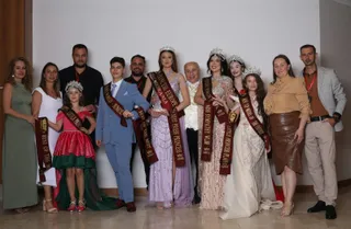 Un grup de persoane posedă panglici decorative și poartă rochii de gală sau costume formale în fața unui perete alb.