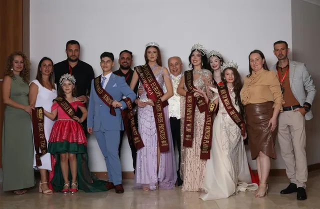 Un grup de persoane posedă panglici decorative și poartă rochii de gală sau costume formale în fața unui perete alb.