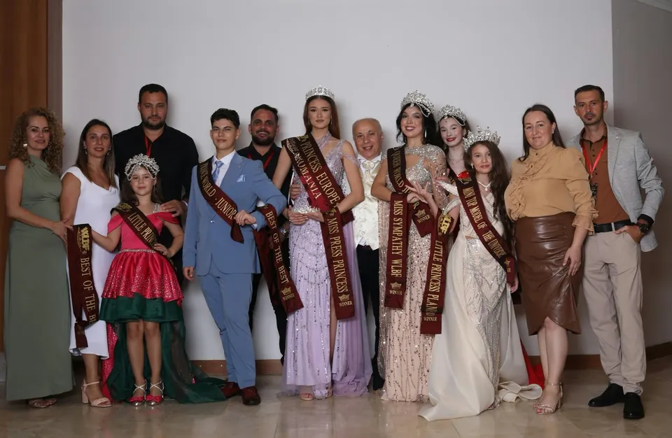 Un grup de persoane posedă panglici decorative și poartă rochii de gală sau costume formale în fața unui perete alb.