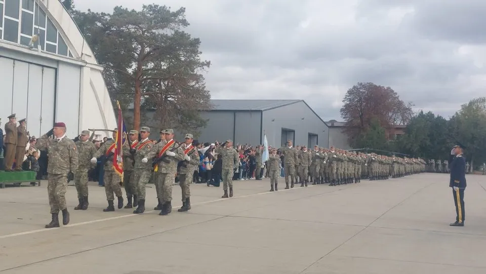 Soldați în uniforme de camuflaj merg în formație pe o platformă de beton lângă un hangar și un grup de oameni.