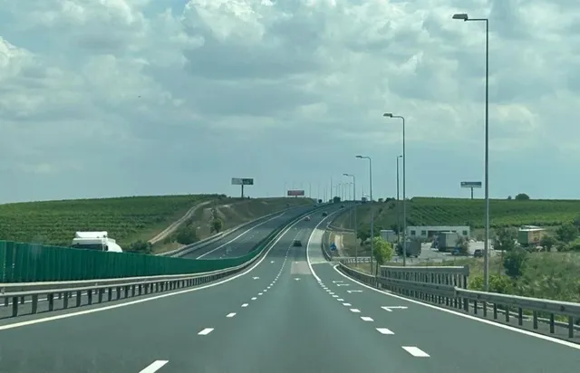 O autostradă cu mai multe benzi de circulație se întinde spre orizont sub un cer înconjurat de nori, fiind deservită de diverse stâlpi de iluminat.