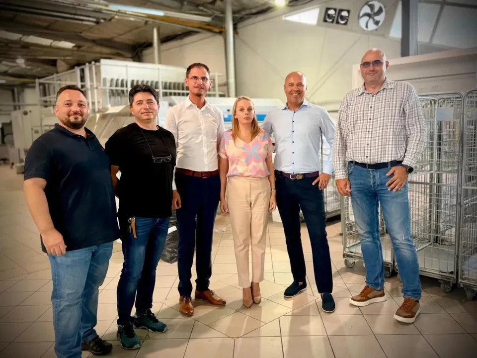 Șase adulți stau aliniați într-o hală, în fața unor containere metalice cu role și a unor instalații industriale.