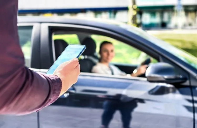 O persoană ține un smartphone în mână, în timp ce un autoturism negru cu un pasager la volan este parcat în fundal.