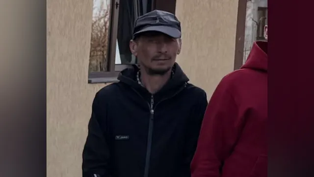 Un bărbat cu șapcă gri și hanorac negru stă în fața unei clădiri lângă o fereastră, fiind parțial acoperit de o persoană în haine roșii.