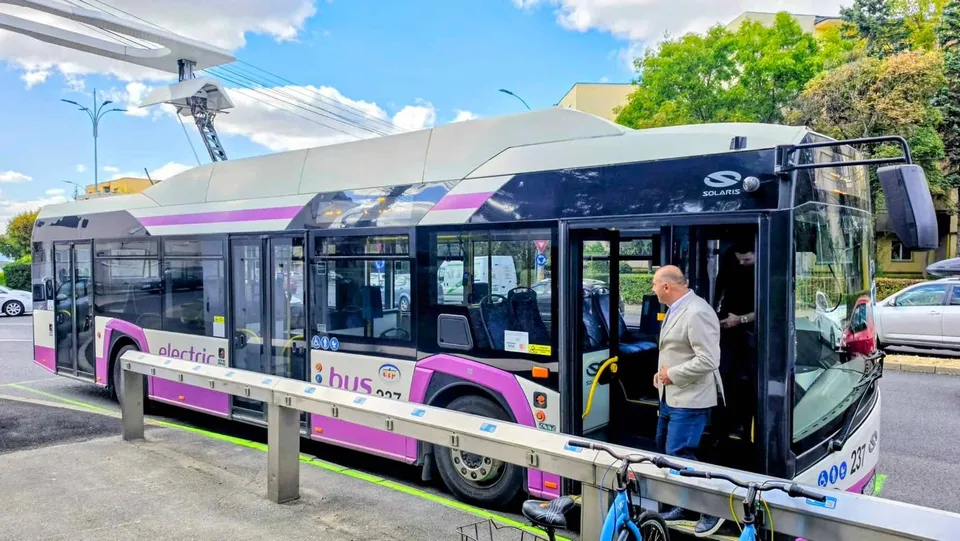 Un autobuz alb cu detalii mov este oprit lângă un gard metalic, având o ușă deschisă prin care intră un bărbat cu sacou.