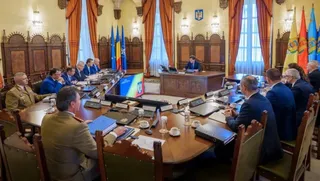 O cameră de ședință cu un masă mare, ovală, în jurul căreia stau mai mulți bărbați în costume și uniforme, cu steaguri în fundal.