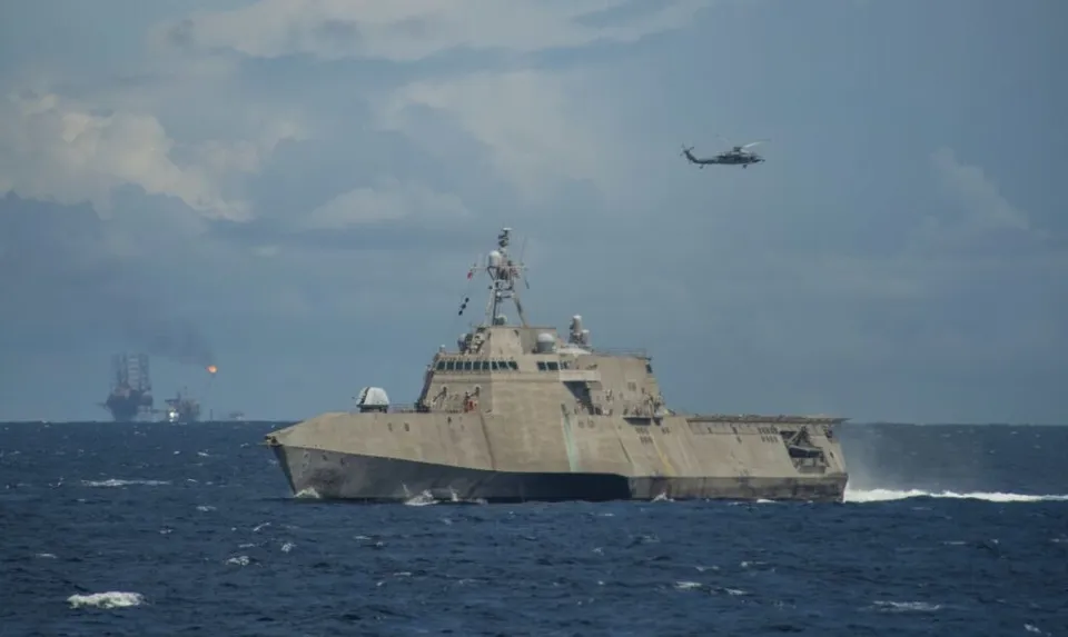 O navă militară gri navigă pe un ocean sub un cer înnorat, în timp ce un elicopter zboară în fundal.