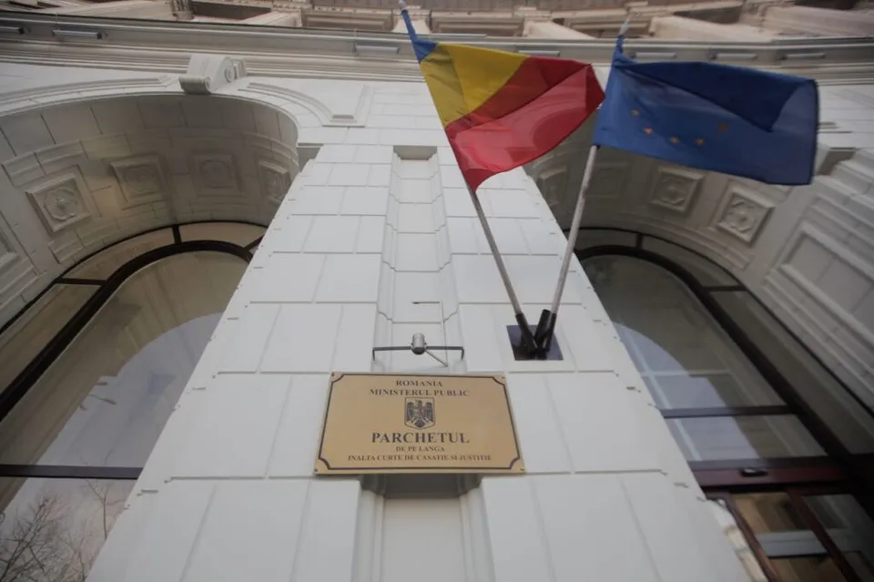 Două steaguri, unul tricolor și unul albastru cu stele galbene, sunt arborate pe fațada unei clădiri albe de lângă o placă cu inscripții.
