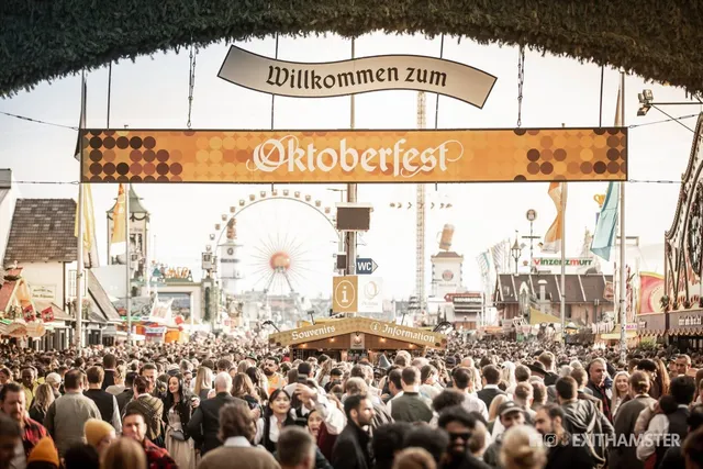 O mulțime de oameni stă în fața unui turn de pâslă cu un panou pe care scrie Oktoberfest, având o roată romantică în fundal.