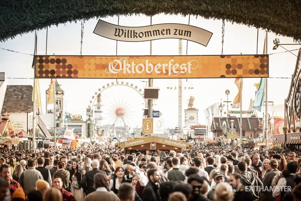 O mulțime de oameni stă în fața unui turn de pâslă cu un panou pe care scrie Oktoberfest, având o roată romantică în fundal.
