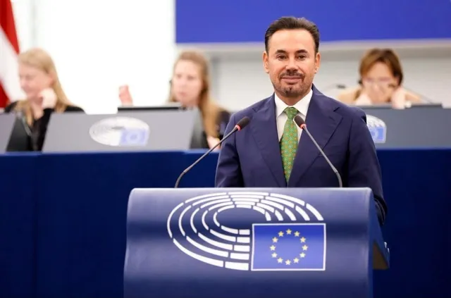 Un bărbat cu barbă și cravată verde stă în spatele unui podium albastru cu emblema Parlamentului European.