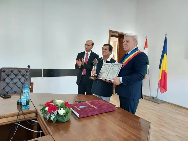 Trei bărbați stau lângă o masă de lemn pe care se află un buchet de flori și un dosar, într-o încăpere cu două steaguri.