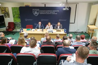 Oamenii stau la mese în fața unui panou albastru pe care sunt scrise texte și simboluri, sub un proiector suspendat.