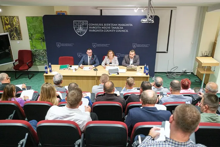 Oamenii stau la mese în fața unui panou albastru pe care sunt scrise texte și simboluri, sub un proiector suspendat.
