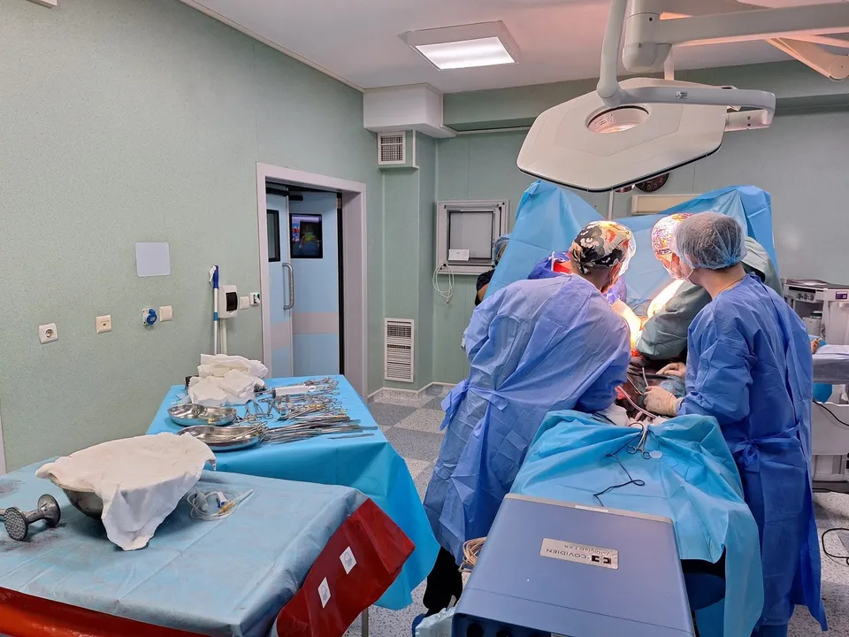 Echipe medicale în halate și mănuși sterile execută o procedură într-o sală de operație lângă mese acoperite cu instrumente metalice și material steril.