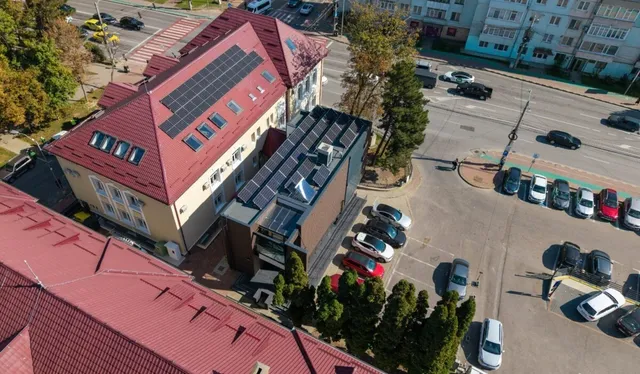 O vedere aeriană prezintă o clădire cu acoperiș roșu dotată cu panouri solare, situată lângă o parcare cu mai multe mașini și drumuri asfaltate.