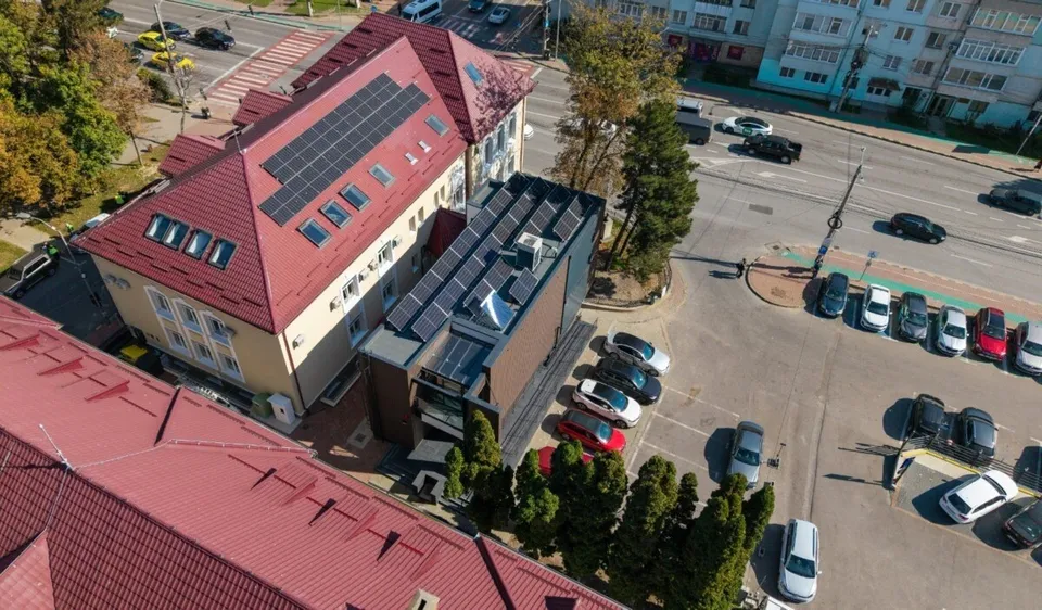 O vedere aeriană prezintă o clădire cu acoperiș roșu dotată cu panouri solare, situată lângă o parcare cu mai multe mașini și drumuri asfaltate.