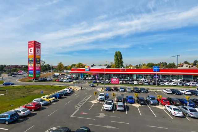 O clădire de retail cu acoperiș roșu este situată lângă un parc de parcare plin de mașini sub un cer senin cu nori subțiri.