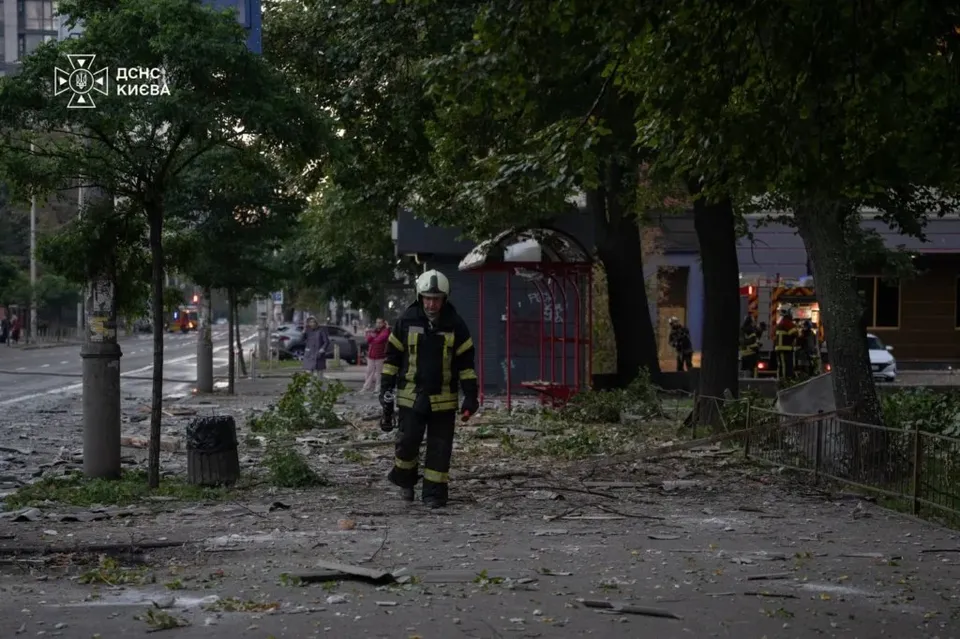 Un bărbat în uniformă de serviciu cu benzi reflectorizante merge pe un trotuar acoperit de resturi și deșeuri lângă un pavilion roșu.