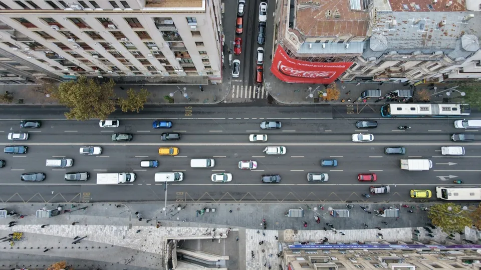 O vedere aeriană de sus a unui bulevard cu mai multe benzi de circulație pe care circulă diverse mașini, autobuze și camioane între clădiri urbane.