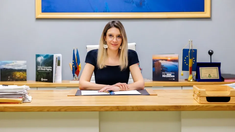 O femeie cu părul blond și ochelari stă la o masă de lemn în fața unui fundal gri cu tablouri și obiecte decorative.