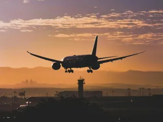 Un avion de pasageri este siluetat în zbor pe un cer la apus, deasupra unei turnițe de control de aeroport.