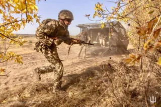 Un soldat în uniformă de camuflaj și cască aleargă prin iarbă uscată ținând un pușcă, în timp ce un vehicul militar este vizibil în fundal.