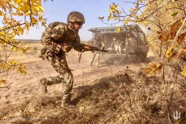 Un soldat în uniformă de camuflaj și cască aleargă prin iarbă uscată ținând un pușcă, în timp ce un vehicul militar se află în fundal.