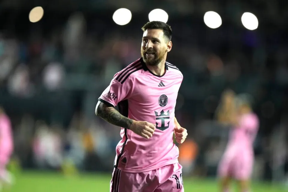 Un bărbat cu barbă, îmbrăcat într-un tricou roz cu dungi negre pe umeri, aleargă pe un teren de fotbal sub lumini de stadion.