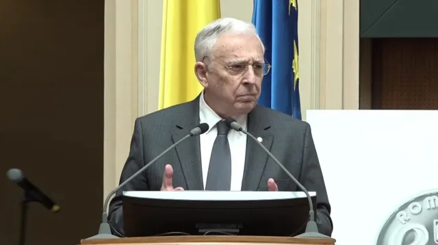 Un bărbat cu ochelari, purtând sacou gri și cravată închisă, stă în spatele unui podium cu microfoane în fața unor steaguri.