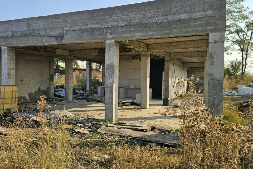 O structură de beton nefinisată cu coloane și tavan plat se află într-un teren cu vegetație uscată și resturi de materiale.
