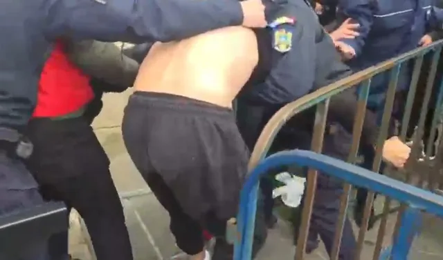 Un bărbat fără tricou este ținut de mai mulți oameni îmbrăcați în uniforme în apropierea unei bariere metalice.