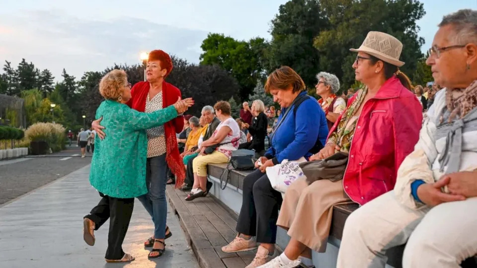 Un grup de persoane stă pe o bancă de lemn în timp ce două femei dansează pe un drum pavat în aer liber.