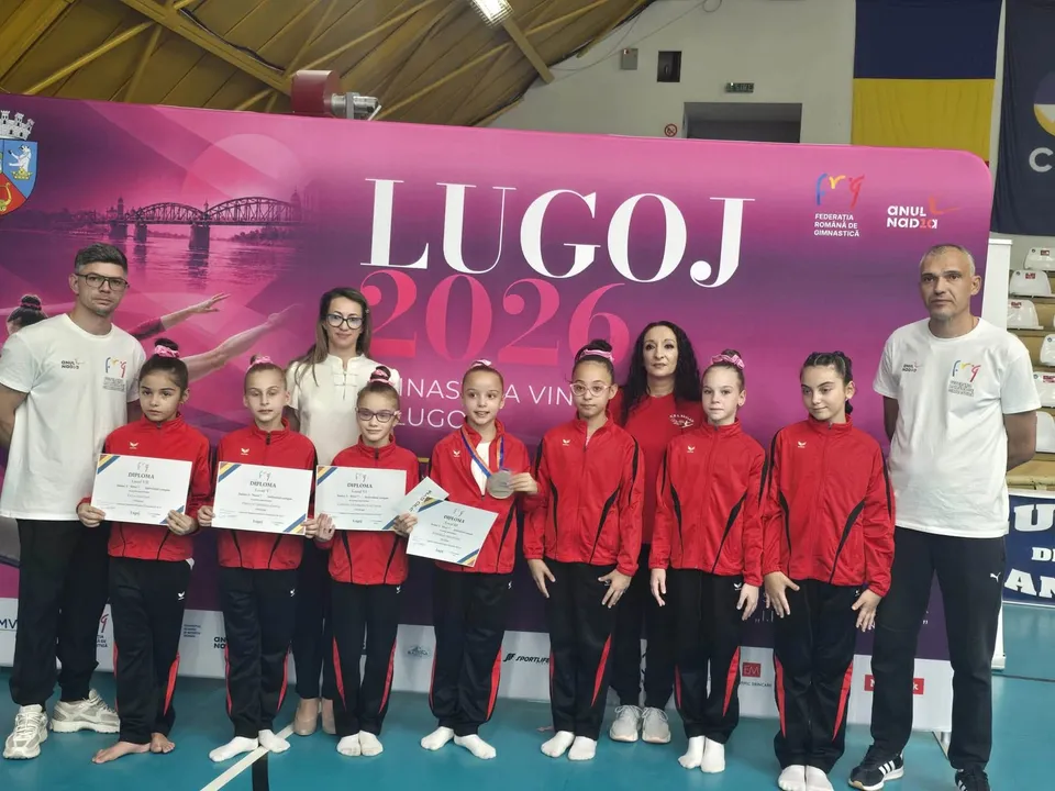 Un grup de gimnaste în echipamente roșii și negre stă în față ținând diplome, alături de patru adulți în fața unui panou roz.