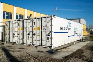 Un container alb mare cu inscripția KLAR100 este amplasat pe o platformă de beton lângă o clădire galbenă sub un cer senin.