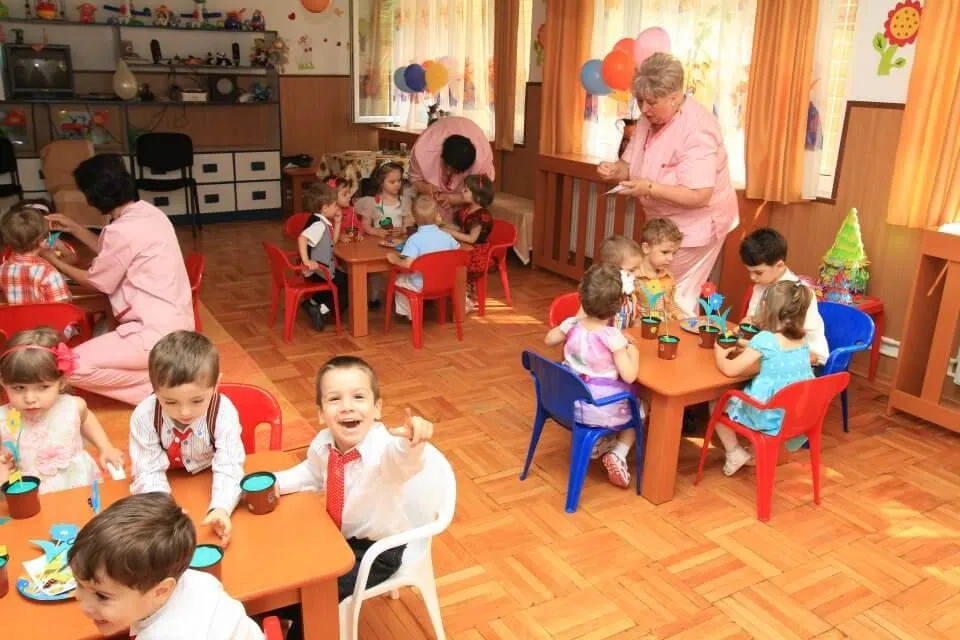 Copii mici stau la mesele de jucărie într-o creșă cu parchet de lemn, însoțiți de adulți în roșu și roz.